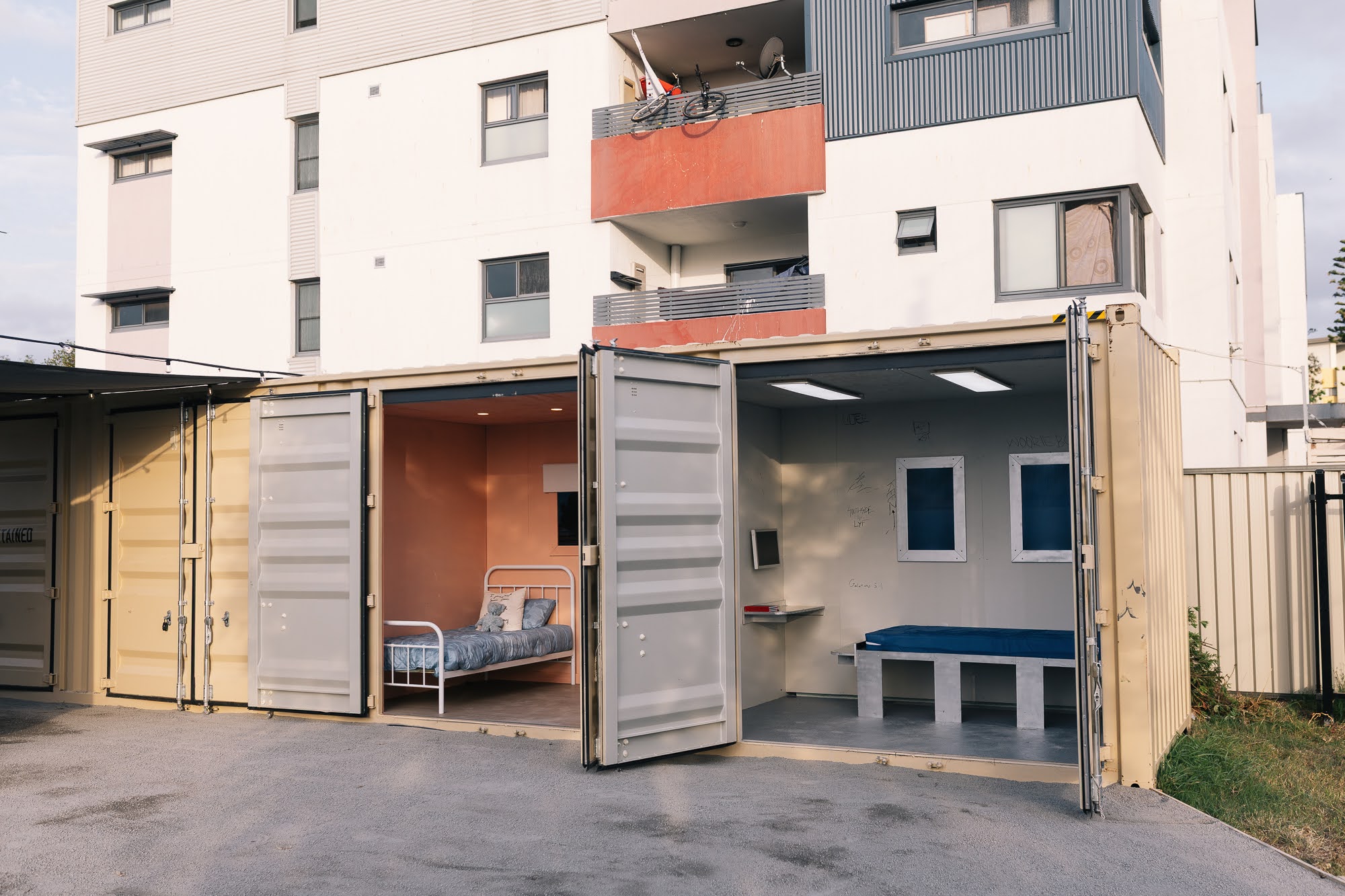 The CONTAINED shipping container in a public courtyard, doors open to a warm bedroom on one side and a bare detention cell on the other