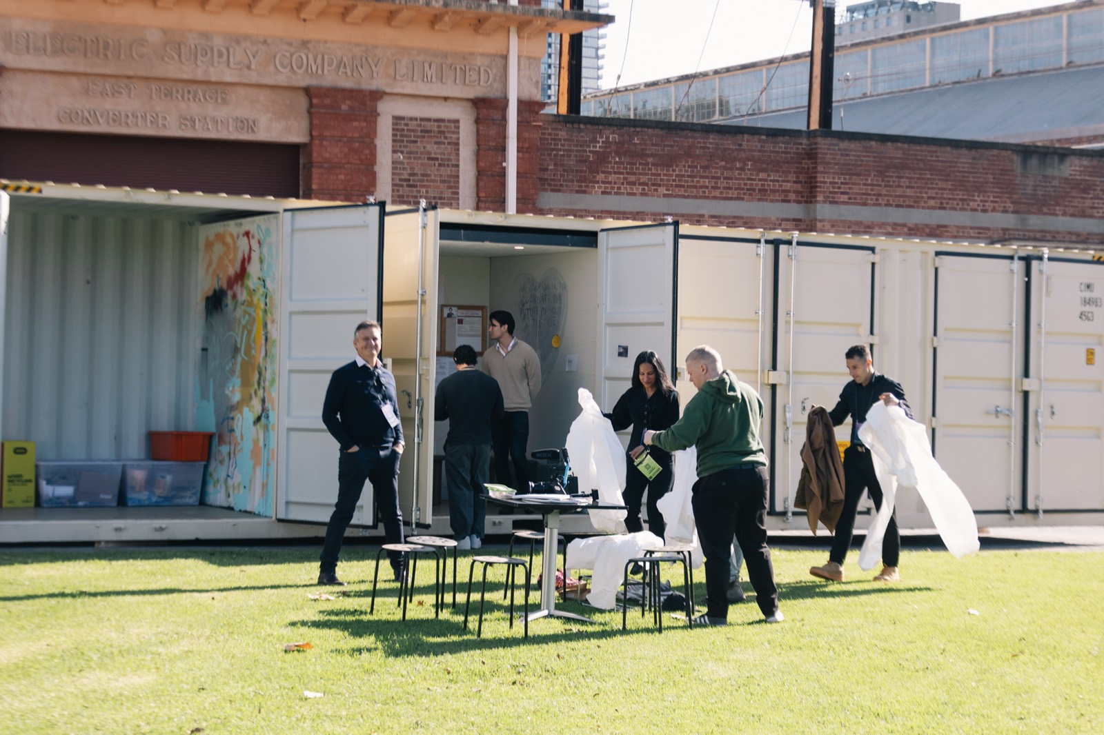 Visitors gathered on the grass outside the open containers in the sun, holding white coveralls, the old Electric Supply Company building behind them