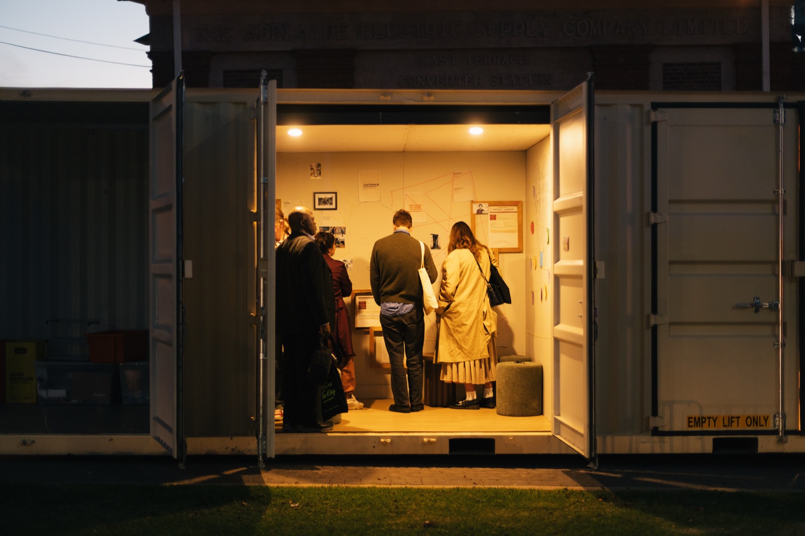 A small group standing close together inside the warmly lit community room at dusk, backs to the street, reading the wall