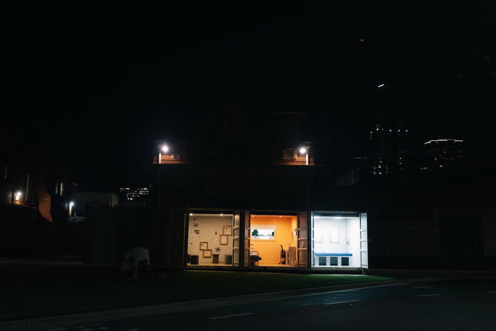 CONTAINED seen from across an empty street late at night, its three rooms lit and open with nobody inside, a construction crane in the dark skyline behind