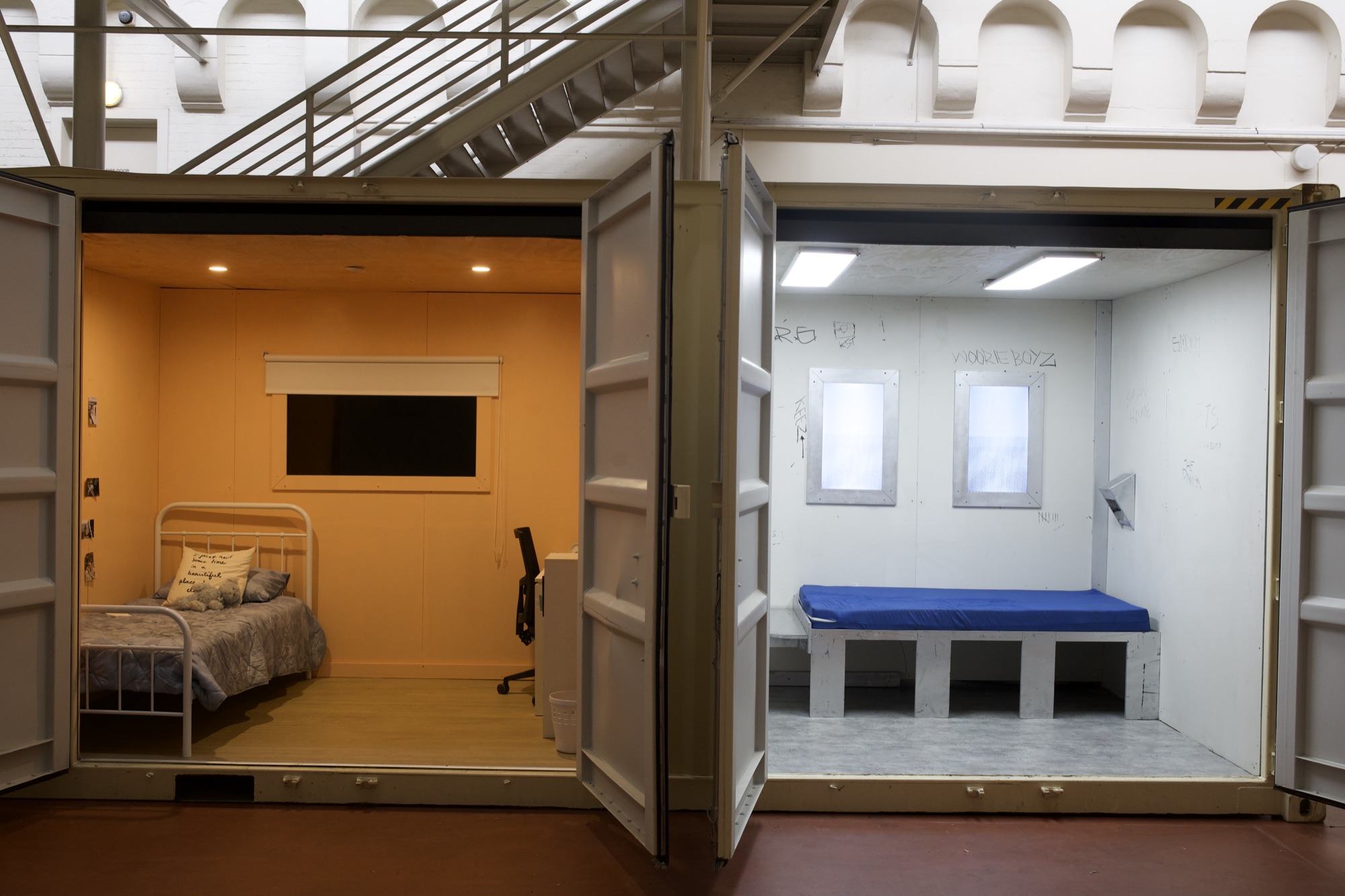 Two finished container rooms side by side: the warm orange bedroom with a made bed and desk, and the white cell with scratched walls and a blue mattress on a concrete plinth