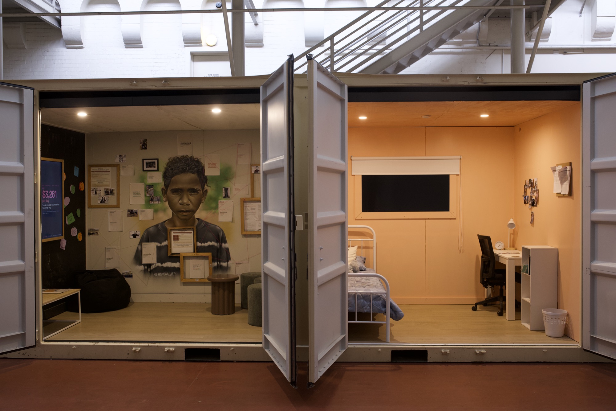 Two finished rooms of CONTAINED side by side inside Tandanya: the community room holding Thomas Readett’s painted portrait of Jackquann surrounded by framed notes, beside the warm orange alternative room with its made bed and desk