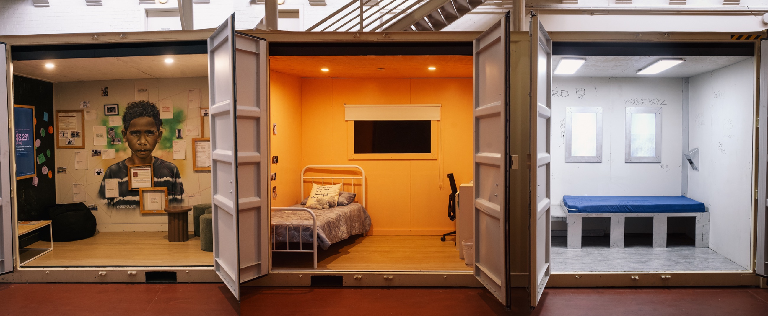 All three finished rooms of CONTAINED side by side inside Tandanya: the community room with Jackquann’s painted portrait, the warm orange alternative room, and the white cell with its blue mattress and scratched walls