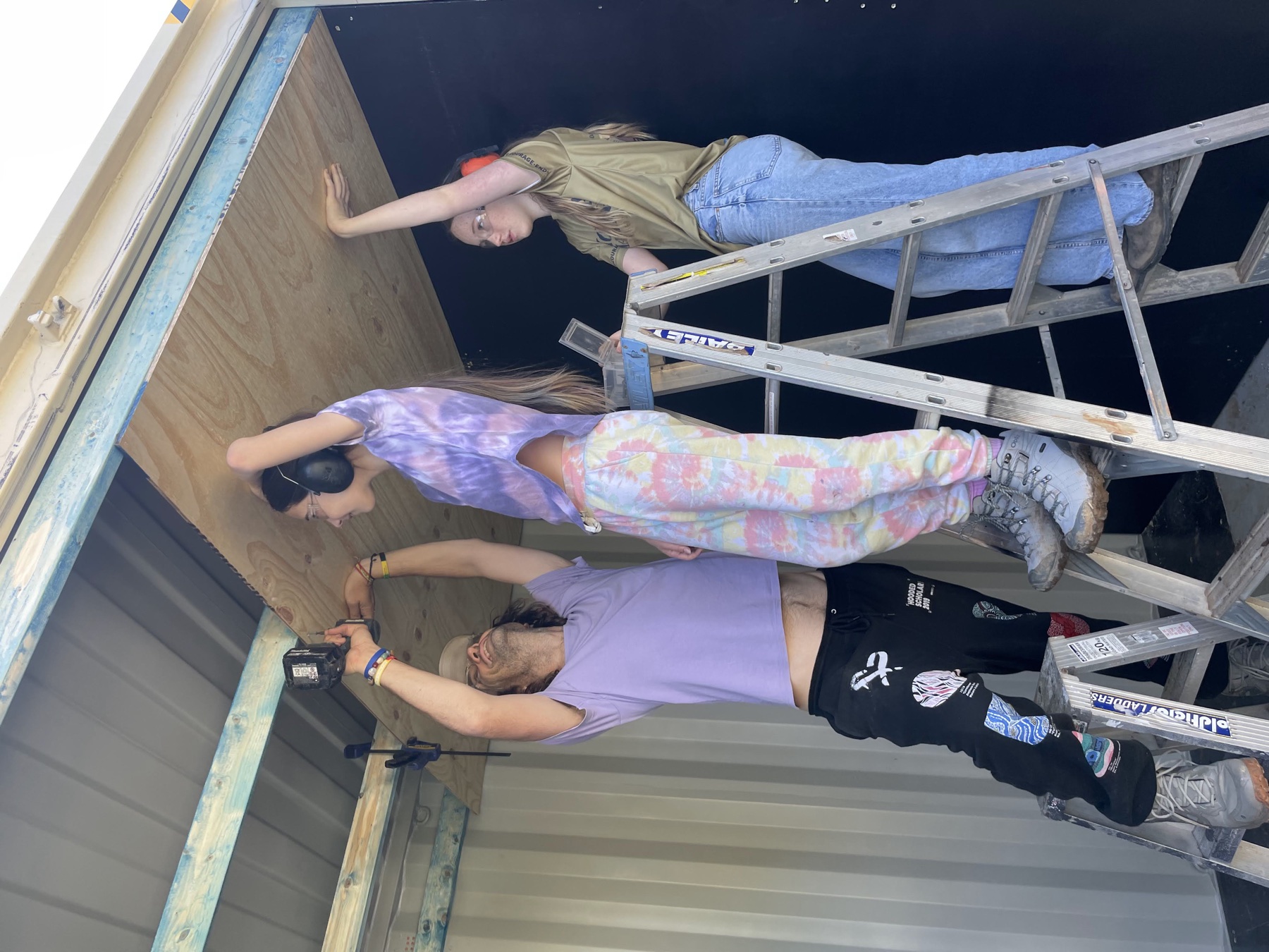 Three young people on ladders inside the container, arms raised, fixing a plywood ceiling panel in place