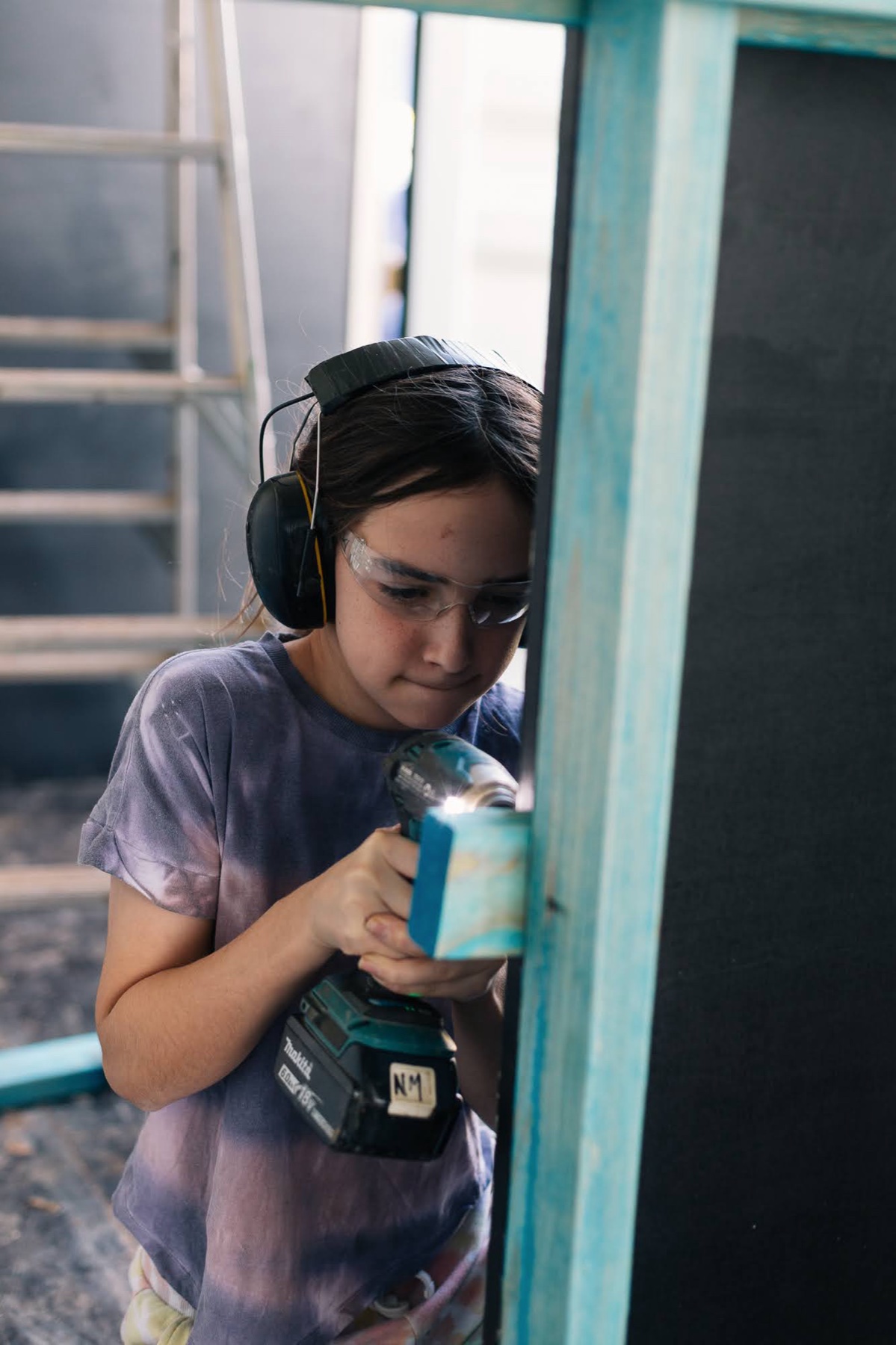 A young girl in ear protection and safety glasses drives a screw into a blue-green timber frame inside the container