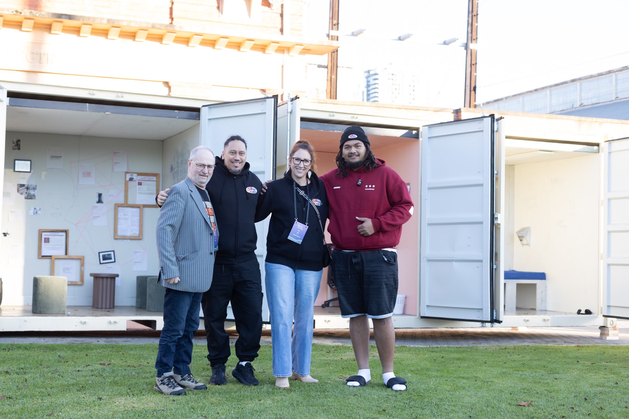 Four people stand arm in arm on the grass in front of the open CONTAINED rooms in Adelaide, Isaiah in a maroon hoodie giving a thumbs up