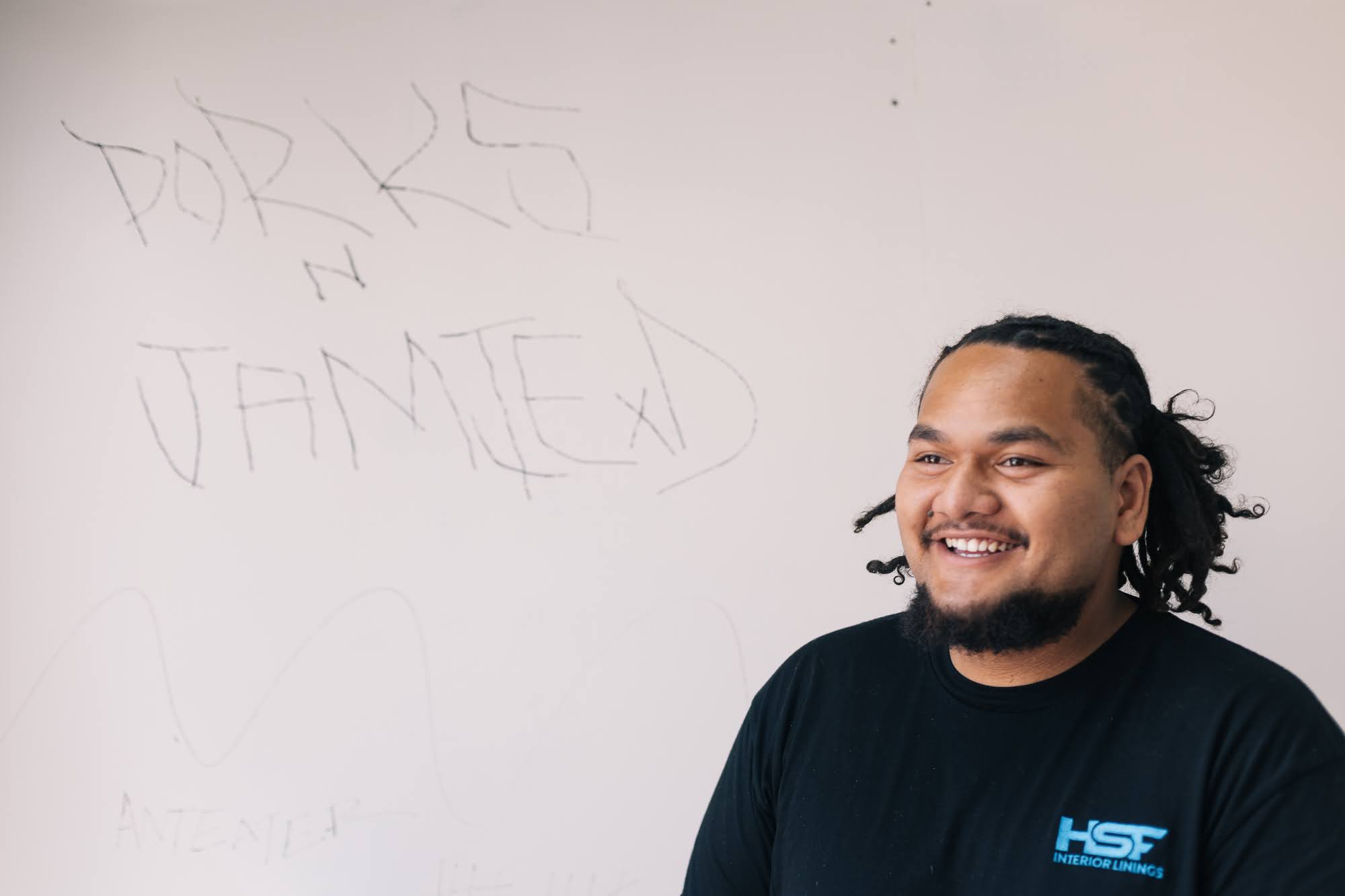 Isaiah, a young man with braided hair, smiles in a CONTAINED room, handwritten wall text reading PORKS N JAMIE behind him