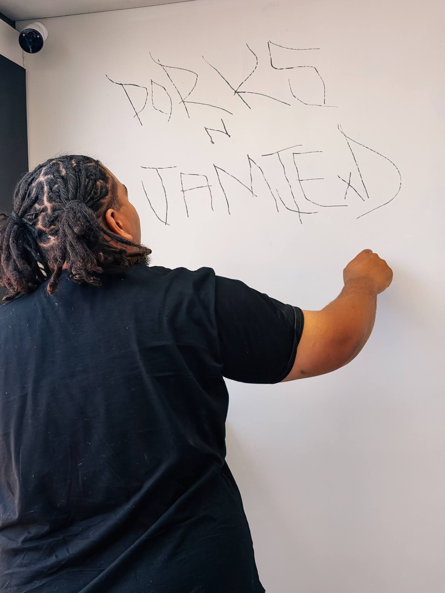Seen from behind, Isaiah writes PORKS N JAMIE on the white wall of the CONTAINED cell room, a camera dome in the corner above him