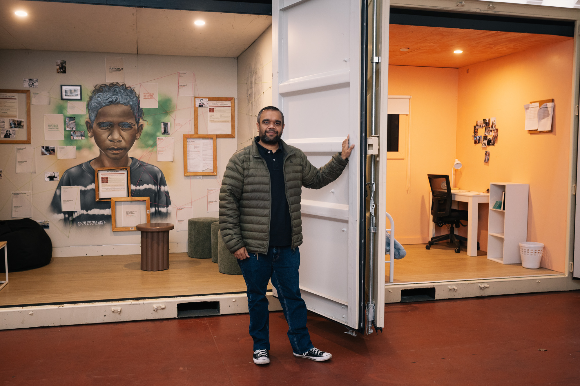 Brenz Saunders stands holding a container door open between the community room, with its painted portrait of Jackquann, and the warm orange alternative room